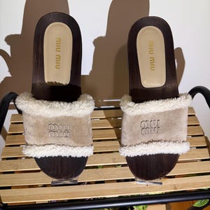 May include: A pair of brown wooden platform sandals with a beige suede strap and white faux fur trim. The sandals have the brand name "miu miu" embossed on the strap. The sandals are displayed on a wooden shelf.