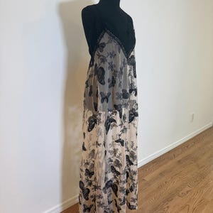 Puede incluir: Un vestido largo y fluido sobre un maniqu&iacute; negro. El vestido presenta un corpi&ntilde;o de encaje negro y una superposici&oacute;n transparente con un estampado de mariposas en negro y gris. El vestido es largo hasta el suelo y tiene un cuello halter.