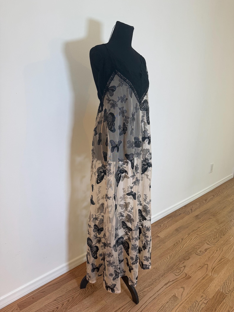 Puede incluir: Un vestido largo y fluido sobre un maniqu&iacute; negro. El vestido presenta un corpi&ntilde;o de encaje negro y una superposici&oacute;n transparente con un estampado de mariposas en negro y gris. El vestido es largo hasta el suelo y tiene un cuello halter.