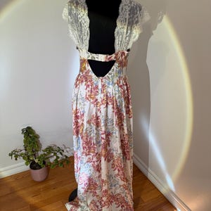 May include: A floral print maxi dress with a white background and pink, yellow, and blue flowers. The dress features a lace back and sleeves. The dress is displayed on a black mannequin.