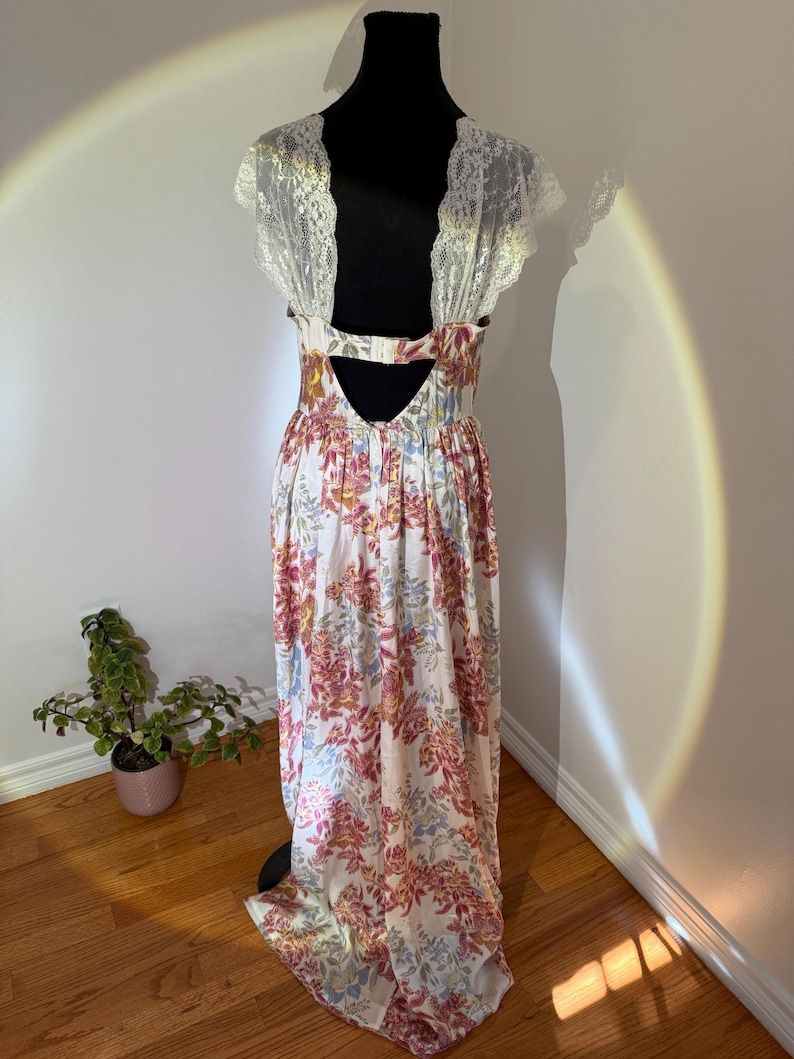 May include: A floral print maxi dress with a white background and pink, yellow, and blue flowers. The dress features a lace back and sleeves. The dress is displayed on a black mannequin.