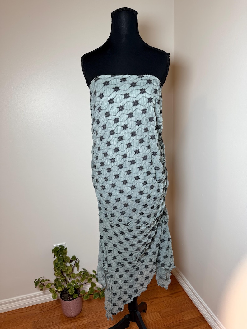 May include: A light blue strapless dress with a black floral pattern, displayed on a black mannequin. The dress features a textured, pleated design and a floor-length, asymmetrical hemline. The dress is made of a lightweight fabric.