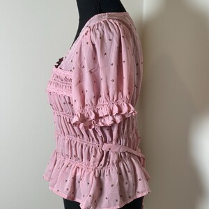 May include: Pink blouse with short, puffed sleeves and a V-neck. The blouse features a shirred bodice, ruffled trim, and a small floral pattern. A tie detail is at the waist. The blouse is displayed on a black mannequin.