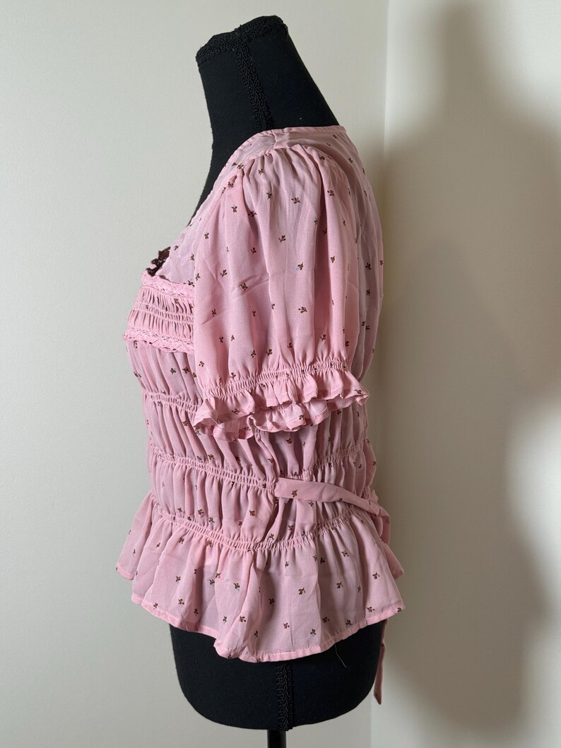 May include: Pink blouse with short, puffed sleeves and a V-neck. The blouse features a shirred bodice, ruffled trim, and a small floral pattern. A tie detail is at the waist. The blouse is displayed on a black mannequin.