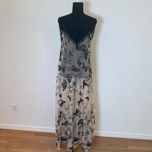 Puede incluir: Un vestido lencero beige con un estampado de mariposas negras. El vestido tiene un escote en V con ribete negro y tirantes finos. Las mariposas est&aacute;n dispersas por todo el vestido, creando un aspecto delicado y aireado. El vestido se exhibe en un maniqu&iacute; negro.
