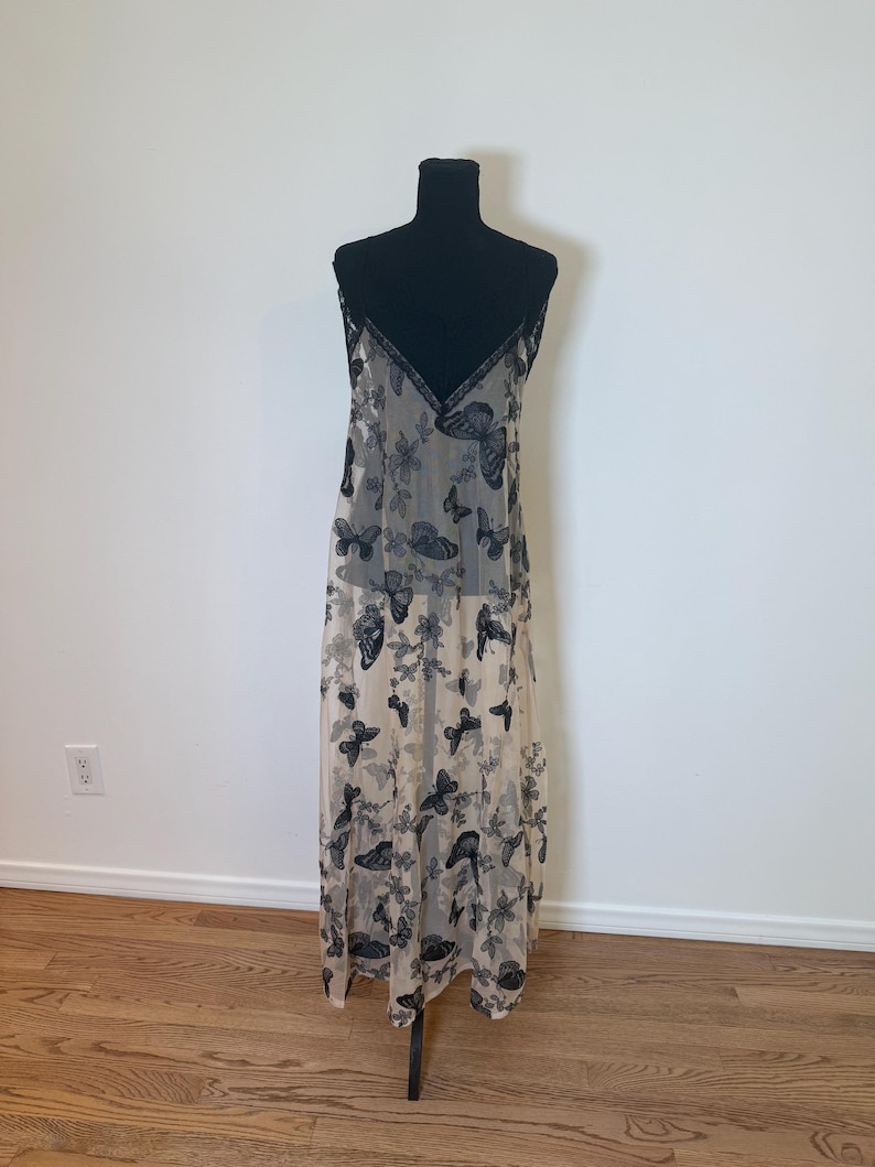 Puede incluir: Un vestido lencero beige con un estampado de mariposas negras. El vestido tiene un escote en V con ribete negro y tirantes finos. Las mariposas est&aacute;n dispersas por todo el vestido, creando un aspecto delicado y aireado. El vestido se exhibe en un maniqu&iacute; negro.