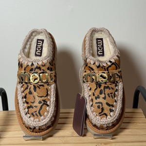 May include: A pair of leopard print slip-on shoes with a tan sole and trim. The shoes have a gold-colored metal accent with the word "mou" on the top. The interior is lined with a cream-colored material. A tag hangs from one shoe.