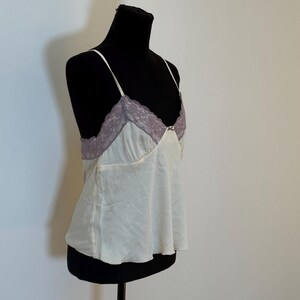 May include: Ivory camisole with thin spaghetti straps and a lilac lace trim. The top is displayed on a black mannequin. The camisole has a flowing, slightly flared shape.