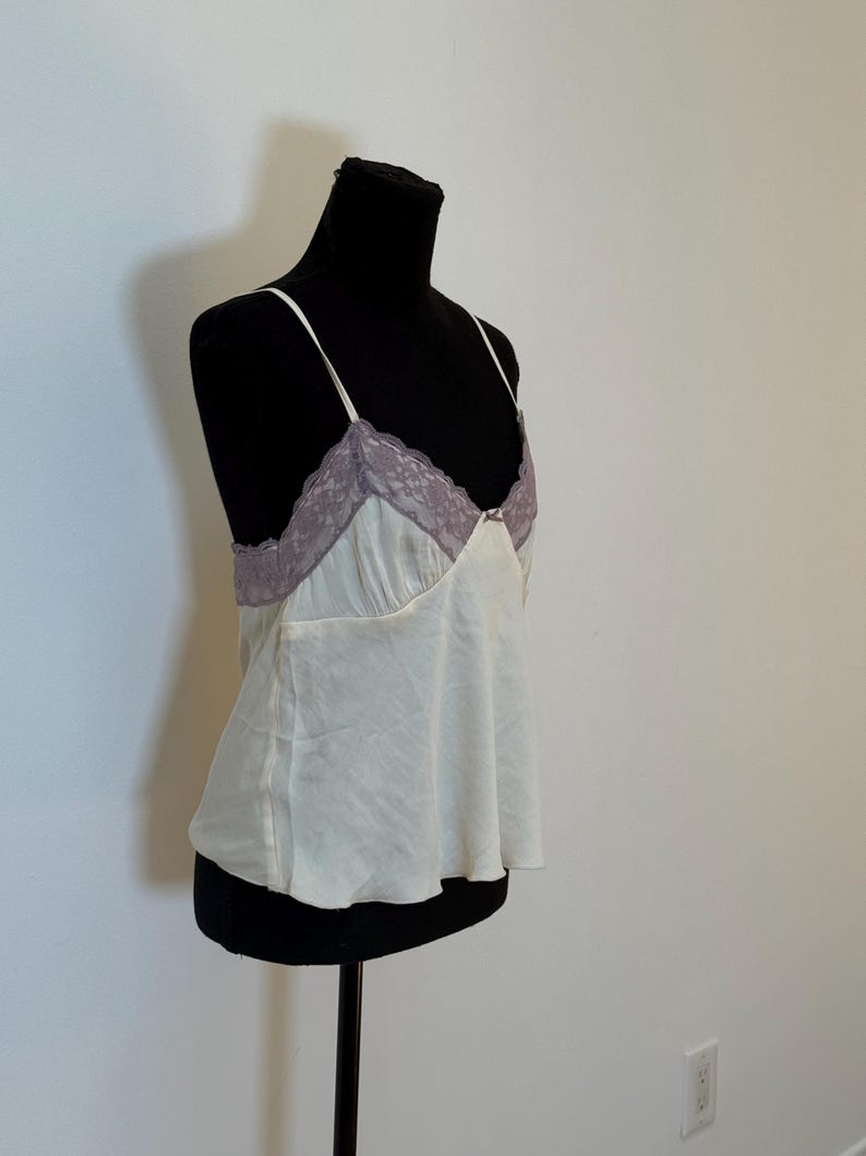 May include: Ivory camisole with thin spaghetti straps and a lilac lace trim. The top is displayed on a black mannequin. The camisole has a flowing, slightly flared shape.
