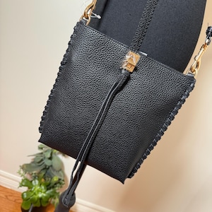 May include: Black crossbody bag with a textured leather appearance. The bag features gold-colored hardware, a long strap, and decorative stitching along the edges. A small tassel hangs from the front. The bag is displayed on a mannequin.
