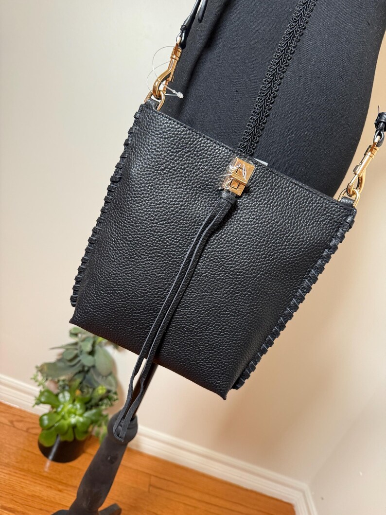 May include: Black crossbody bag with a textured leather appearance. The bag features gold-colored hardware, a long strap, and decorative stitching along the edges. A small tassel hangs from the front. The bag is displayed on a mannequin.