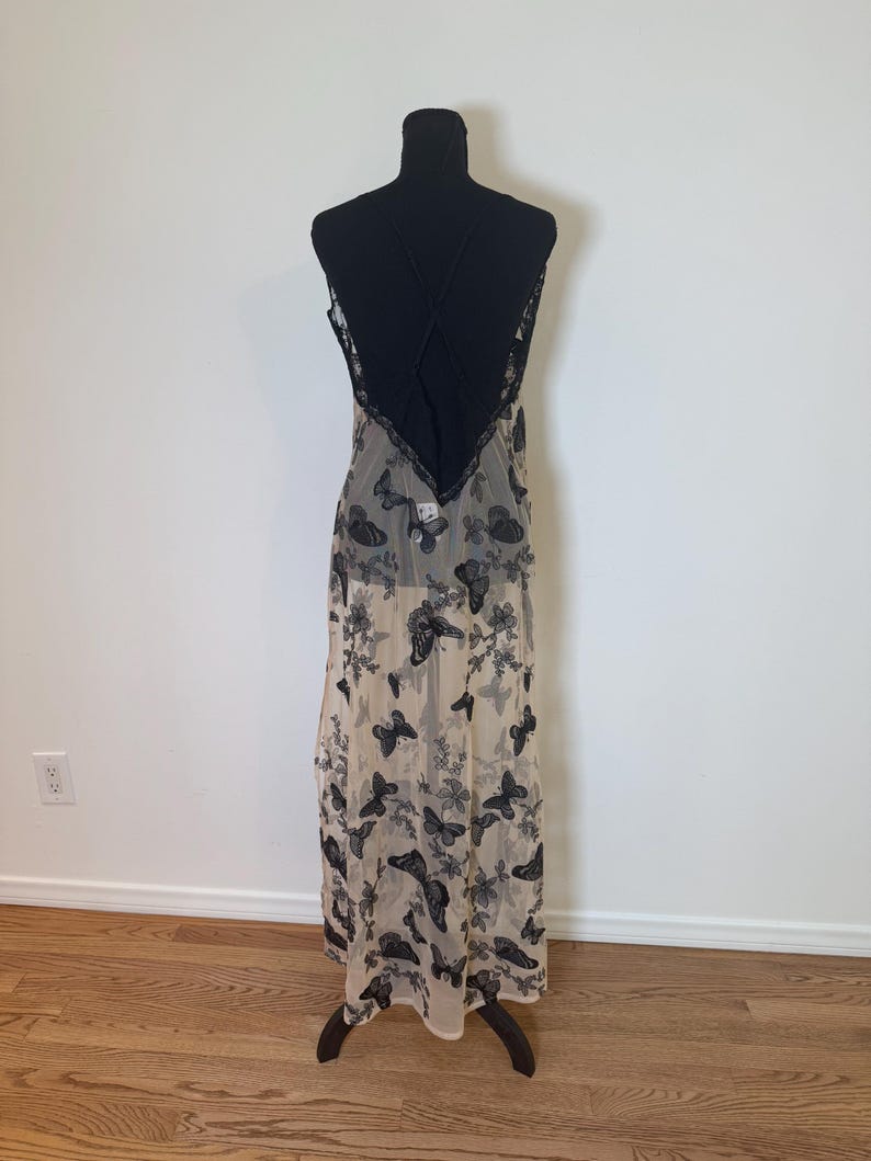 Puede incluir: Un vestido largo y transparente con estampado de mariposas. El vestido presenta un corpi&ntilde;o negro con espalda cruzada y ribete de encaje. La falda es de color beige claro con dise&ntilde;os de mariposas y flores negras.