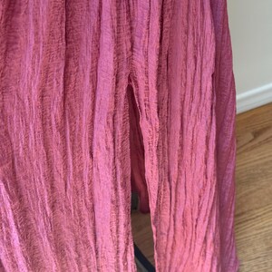 May include: Close-up of a flowing, floor-length dress in a deep rose colour. The fabric has a crinkled texture, with vertical folds creating a draped effect. A slit reveals a glimpse of the legs. The dress appears to be made of a lightweight material.