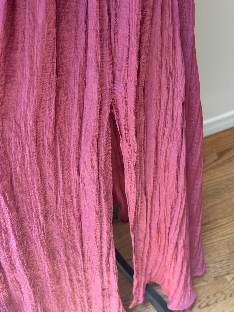 May include: Close-up of a flowing, floor-length dress in a deep rose colour. The fabric has a crinkled texture, with vertical folds creating a draped effect. A slit reveals a glimpse of the legs. The dress appears to be made of a lightweight material.