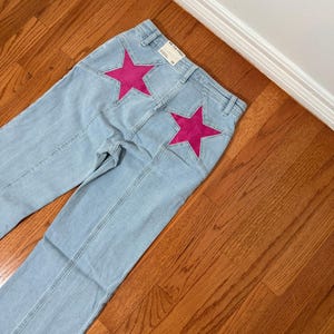Puede incluir: Vaqueros de mezclilla azul claro con dos grandes estrellas rosas fucsia en los bolsillos traseros. Los vaqueros tienen un dise&ntilde;o cl&aacute;sico de cinco bolsillos y corte recto. La etiqueta dice "WE THE FREE".
