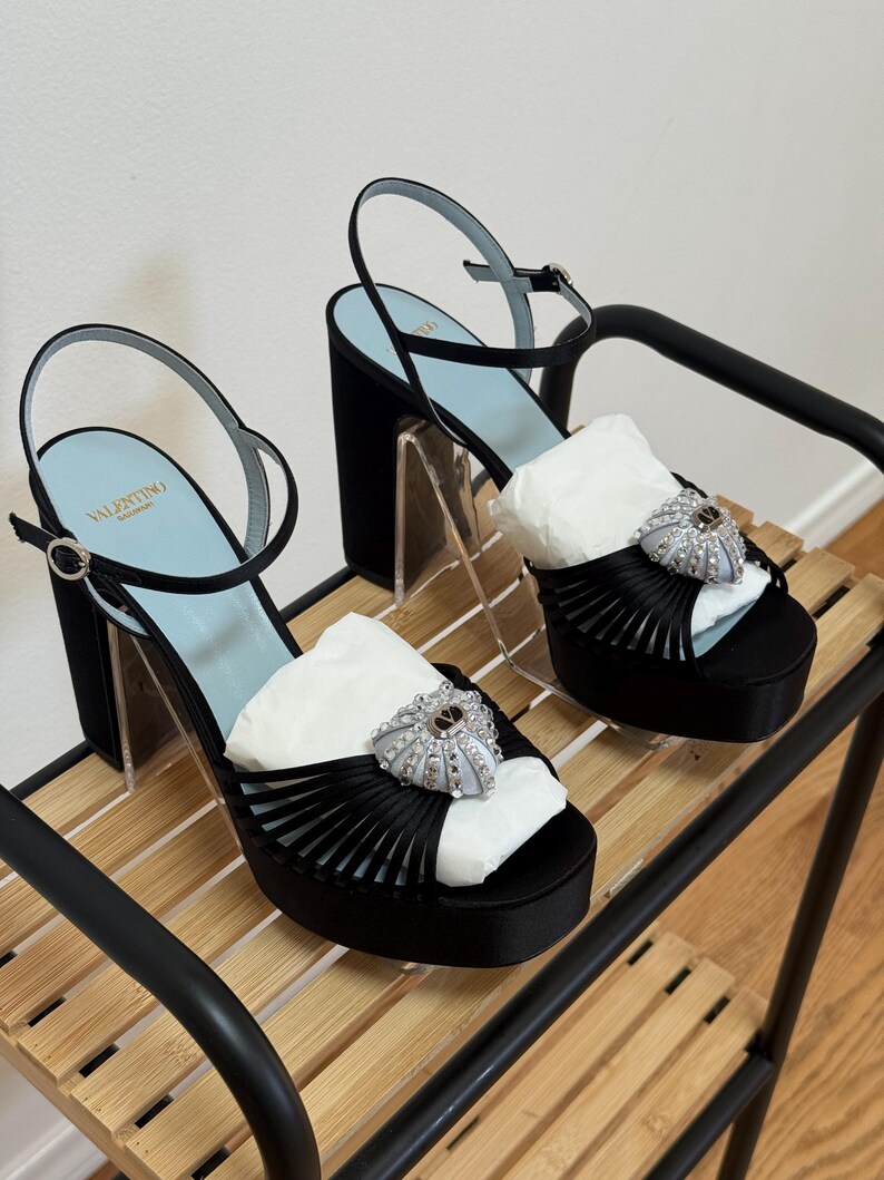 May include: A pair of black satin platform sandals with high block heels and ankle straps. The shoes feature a light blue insole with the Valentino Garavani logo and a decorative silver-toned heart-shaped embellishment with crystals on the front.