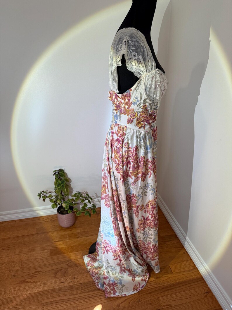 May include: A vintage-style floral print dress on a black mannequin. The dress features a white base with a colorful floral pattern in shades of pink, orange, and blue. The bodice has lace detailing, and the dress appears to be floor-length.