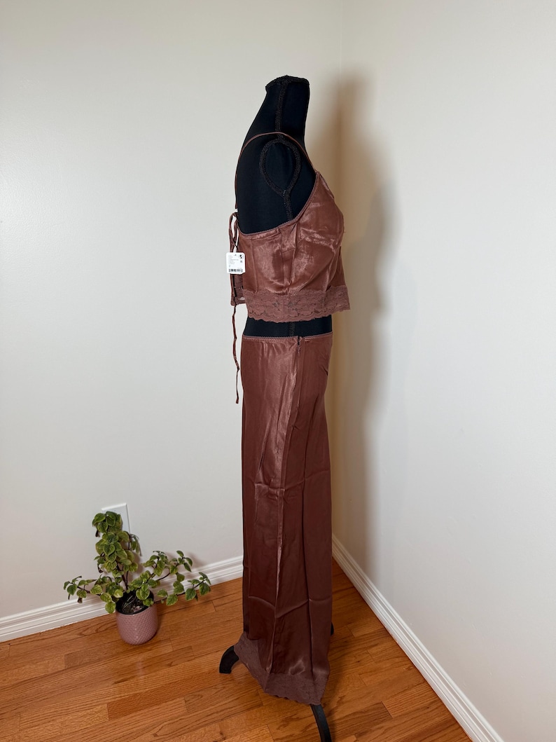 Puede incluir: Conjunto de dos piezas de sat&eacute;n marr&oacute;n sobre un maniqu&iacute;. La parte superior es un camisola con ribete de encaje, y la falda es larga, de cintura alta. El conjunto es de color liso y tiene una etiqueta.
