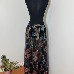 以下が含まれることがあります： マネキンに展示された、花柄の黒いマキシスカート。 スカートは、ウエストバンドにカラフルな花とスパンコールで飾られたシアーオーバーレイが特徴です。 スカートは長く流れるようなデザインで、さまざまな場面に適しています。