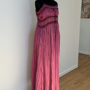 May include: A long, flowing pink dress with a textured, pleated design. The dress features a fitted bodice with black trim and spaghetti straps. The dress is displayed on a black mannequin.