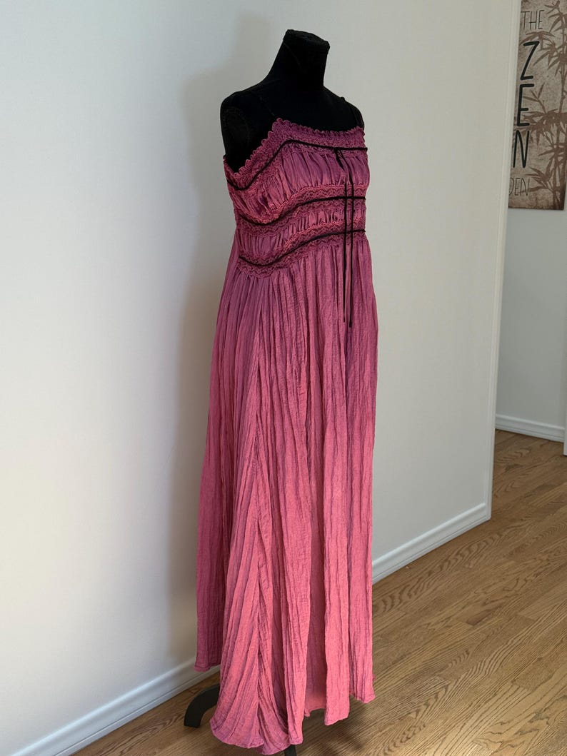 May include: A long, flowing pink dress with a textured, pleated design. The dress features a fitted bodice with black trim and spaghetti straps. The dress is displayed on a black mannequin.
