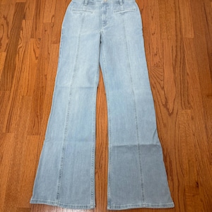 Puede incluir: Pantalones vaqueros azul claro con cintura alta y perneras acampanadas. Los vaqueros tienen costuras verticales en la parte delantera y trasera de las perneras. Los pantalones son de un tejido vaquero de lavado claro.