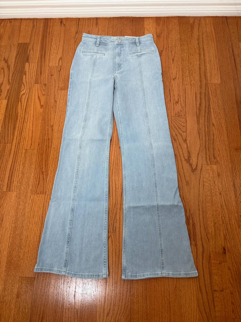 Puede incluir: Pantalones vaqueros azul claro con cintura alta y perneras acampanadas. Los vaqueros tienen costuras verticales en la parte delantera y trasera de las perneras. Los pantalones son de un tejido vaquero de lavado claro.