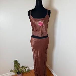 Puede incluir: Conjunto de dos piezas de sat&eacute;n marr&oacute;n sobre un maniqu&iacute;. La parte superior es un camisola con escote cuadrado y un bordado de rosa rosa. La parte inferior es una falda larga. El conjunto se exhibe contra una pared blanca.
