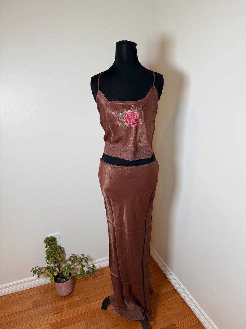 Puede incluir: Conjunto de dos piezas de sat&eacute;n marr&oacute;n sobre un maniqu&iacute;. La parte superior es un camisola con escote cuadrado y un bordado de rosa rosa. La parte inferior es una falda larga. El conjunto se exhibe contra una pared blanca.