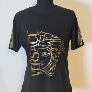 May include: A black short-sleeved t-shirt featuring a large gold metallic graphic of the Versace Medusa head logo and the word VERSACE printed vertically. The sleeves are accented with a gold patterned trim featuring the brand name.