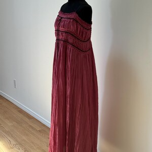 May include: A long, flowing, burgundy dress with thin straps and a gathered bodice. The dress features black trim and a textured fabric, displayed on a black mannequin. The dress is floor-length and suitable for a variety of occasions.