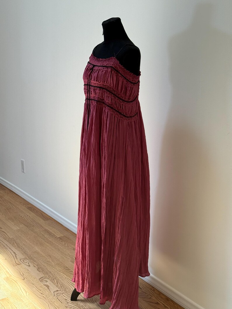 May include: A long, flowing, burgundy dress with thin straps and a gathered bodice. The dress features black trim and a textured fabric, displayed on a black mannequin. The dress is floor-length and suitable for a variety of occasions.