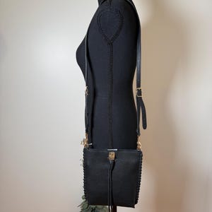 May include: Black crossbody bag with a textured finish, displayed on a black mannequin. The bag features a long, adjustable strap, gold-colored hardware, and a tassel detail. The bag is approximately 8 inches wide and 9 inches tall.