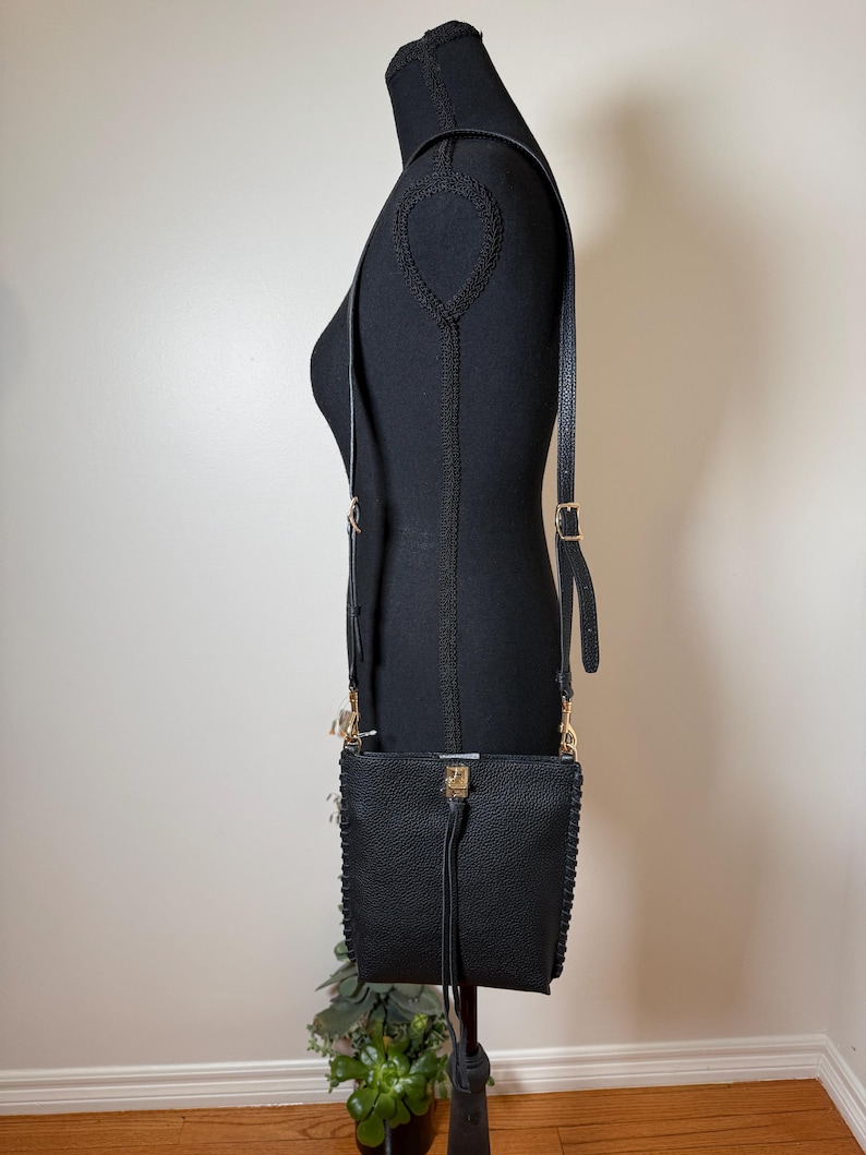 May include: Black crossbody bag with a textured finish, displayed on a black mannequin. The bag features a long, adjustable strap, gold-colored hardware, and a tassel detail. The bag is approximately 8 inches wide and 9 inches tall.