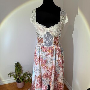 May include: A floral print dress with a white lace bodice and sleeves is displayed on a black mannequin. The dress features a white background with a pattern of pink, yellow, and blue flowers. The skirt has a split front.