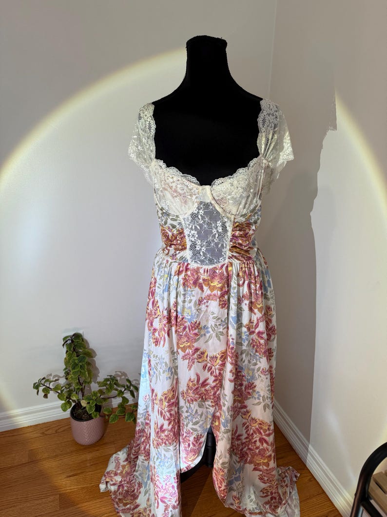 May include: A floral print dress with a white lace bodice and sleeves is displayed on a black mannequin. The dress features a white background with a pattern of pink, yellow, and blue flowers. The skirt has a split front.