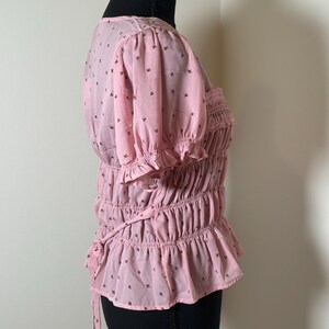 May include: A light pink blouse with short, puffed sleeves and a ruffled hem. The blouse features a smocked bodice and is adorned with small, dark brown floral patterns. A tie detail is visible at the waist.