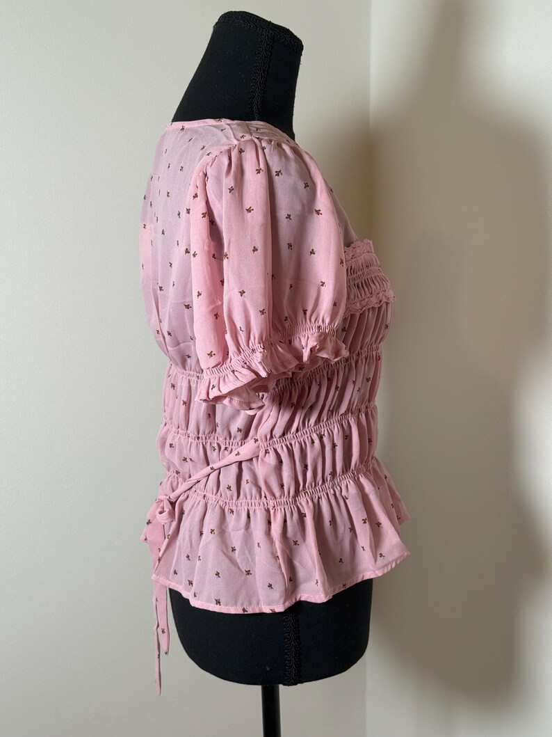 May include: A light pink blouse with short, puffed sleeves and a ruffled hem. The blouse features a smocked bodice and is adorned with small, dark brown floral patterns. A tie detail is visible at the waist.