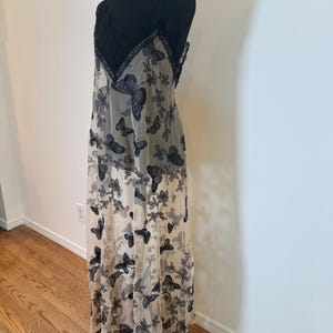 Puede incluir: Un vestido largo y transparente con corpi&ntilde;o de encaje negro y tirantes finos. El vestido presenta una falda color crema adornada con un estampado de mariposas negras y dise&ntilde;os florales. El vestido se exhibe en un maniqu&iacute; negro.