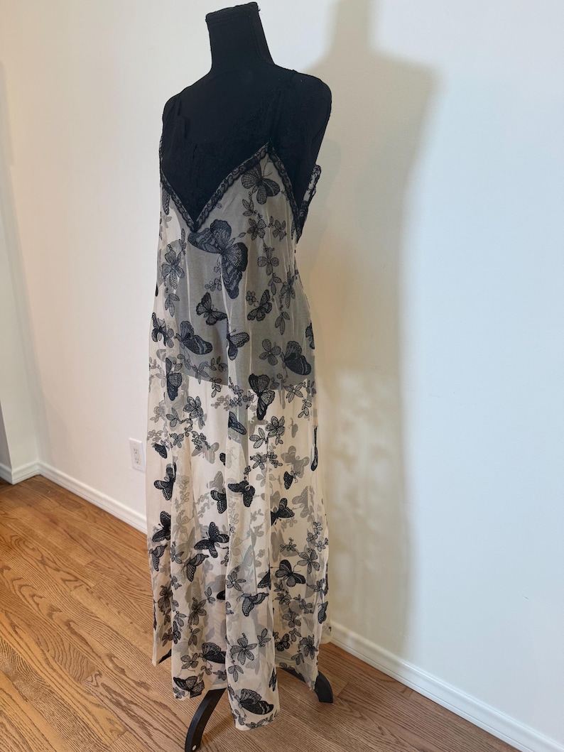 Puede incluir: Un vestido largo y transparente con corpi&ntilde;o de encaje negro y tirantes finos. El vestido presenta una falda color crema adornada con un estampado de mariposas negras y dise&ntilde;os florales. El vestido se exhibe en un maniqu&iacute; negro.