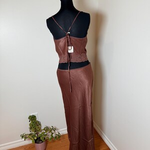 Puede incluir: Conjunto de vestido de sat&eacute;n marr&oacute;n de dos piezas sobre un maniqu&iacute; negro. La parte superior es un camisola con tirantes finos y detalle de encaje en la cintura. La falda es larga, fluida, con ribete de encaje en el dobladillo. Una etiqueta est&aacute; adjunta al vestido.