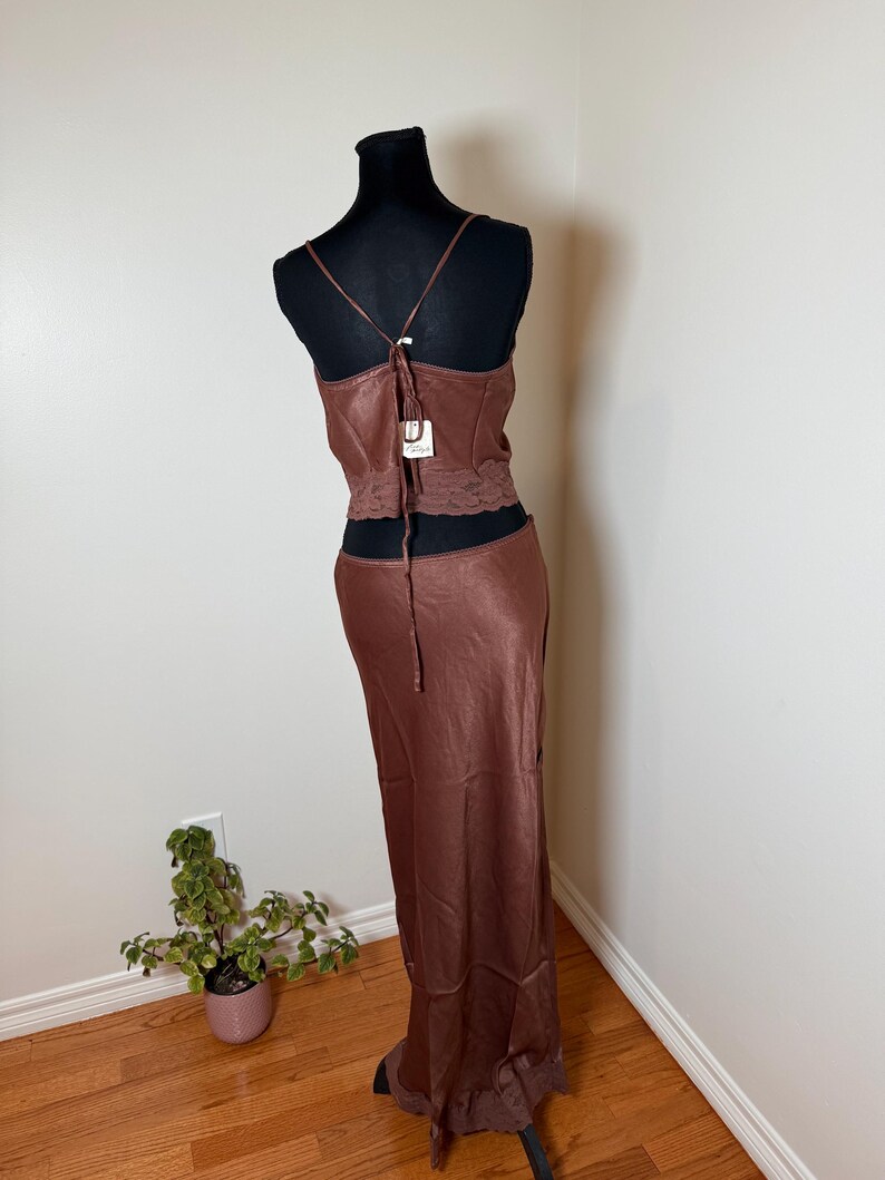 Puede incluir: Conjunto de vestido de sat&eacute;n marr&oacute;n de dos piezas sobre un maniqu&iacute; negro. La parte superior es un camisola con tirantes finos y detalle de encaje en la cintura. La falda es larga, fluida, con ribete de encaje en el dobladillo. Una etiqueta est&aacute; adjunta al vestido.