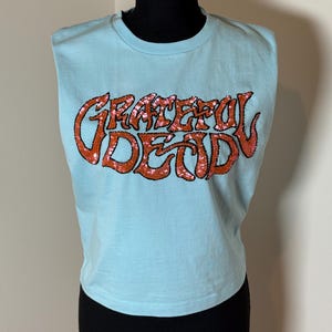 May include: Light blue sleeveless t-shirt with the words "Grateful Dead" in orange sequined lettering. The shirt is displayed on a black mannequin. The text is in a stylized, psychedelic font.