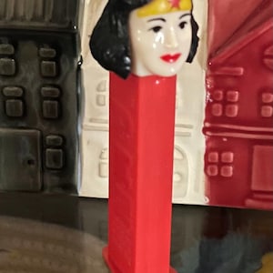 May include: A red PEZ dispenser featuring the head of Wonder Woman. The head is white with black hair, a gold tiara with a red star, and red lipstick. The dispenser is red and has a rectangular shape.