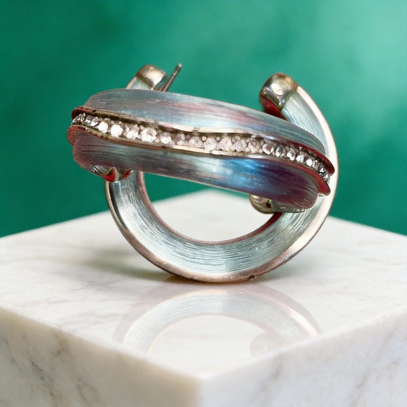 May include: A silver-toned hoop earring with a blue and red enamel design. The earring features a row of small, clear crystals. The earring is set against a green background and rests on a white marble surface.