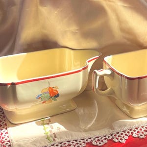 Puede incluir: Dos cuencos de cerámica vintage de color crema con ribete rojo y diseño de frutas. Cada cuenco tiene un asa y un pico. Se colocan sobre un mantel individual blanco con una superficie roja en el fondo.