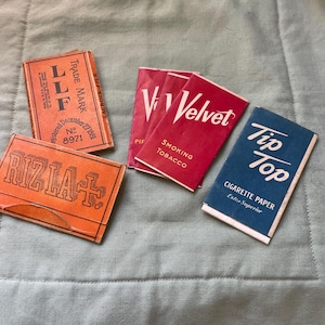 May include: An assortment of vintage cigarette and tobacco packaging. The image features packs of "Velvet" smoking tobacco, "Tip Top" cigarette paper, "LLF" and "Rizla+" brands, all in various colors like red, orange, and blue.