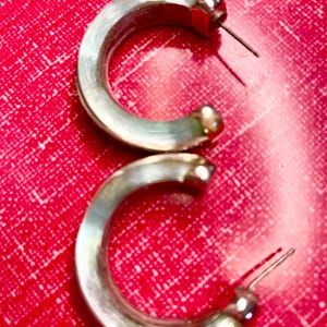 May include: A pair of silver-coloured hoop earrings. The earrings have a rounded, C-shaped design with a post and a small, round accent at the end. The background is a textured, bright red surface.