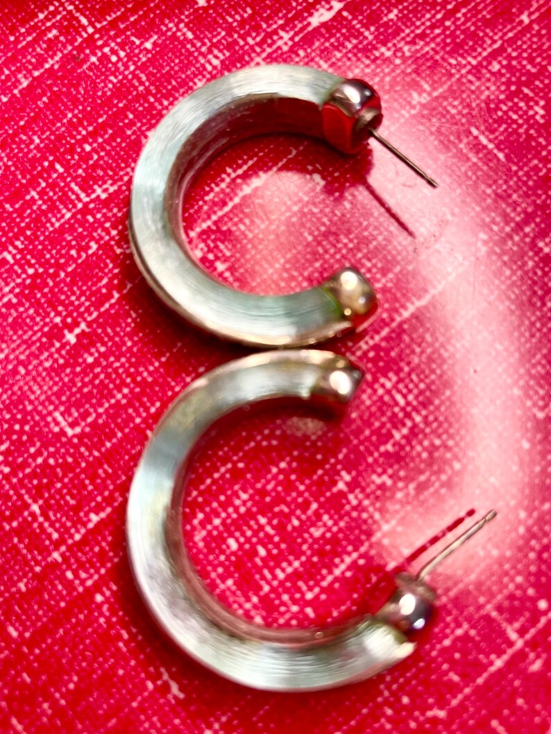 May include: A pair of silver-coloured hoop earrings. The earrings have a rounded, C-shaped design with a post and a small, round accent at the end. The background is a textured, bright red surface.