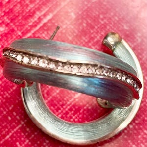 May include: A silver hoop earring with a blue and silver enamel finish. The earring has a row of small, clear crystals. The background is a bright red. The earring is a stylish accessory.